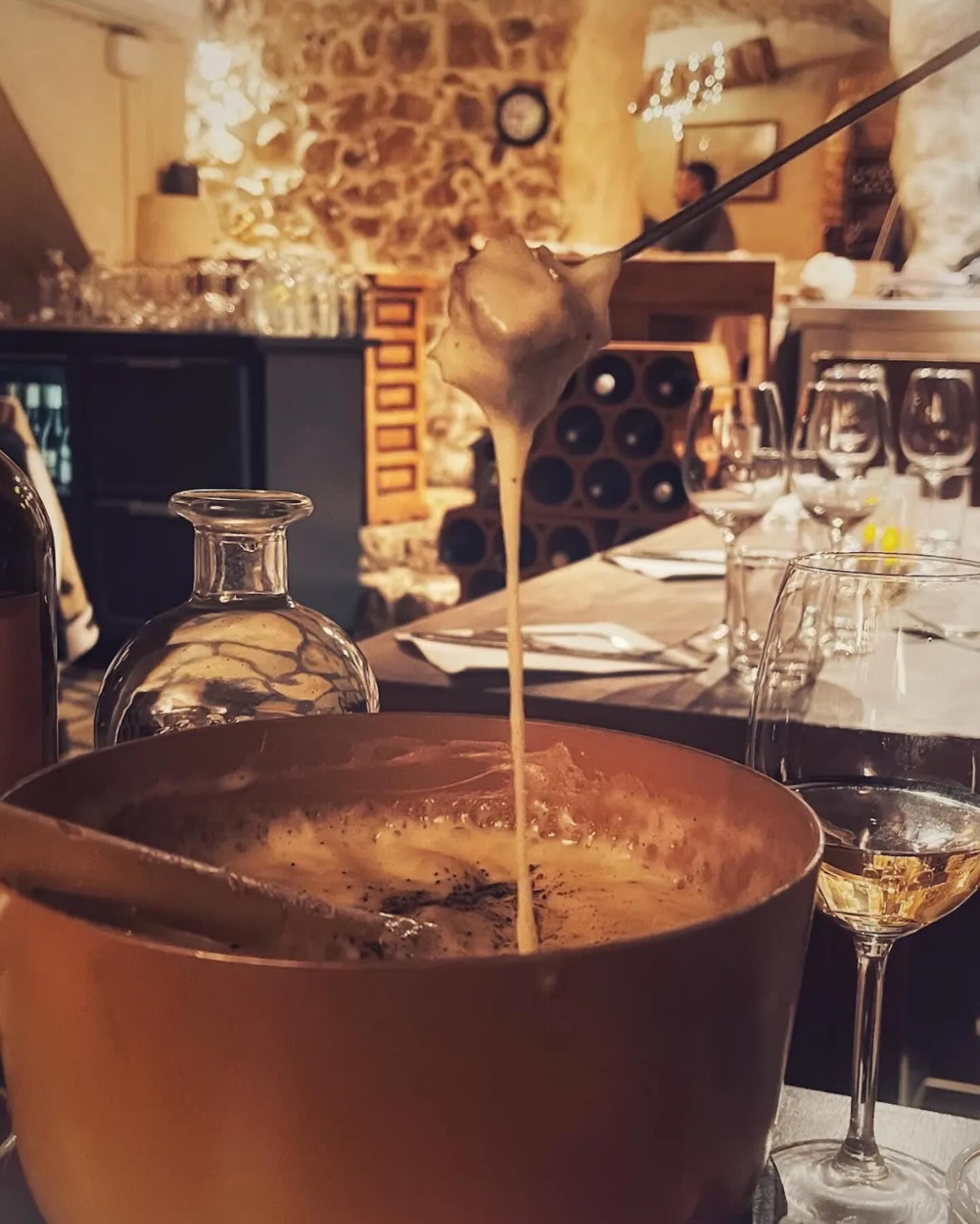 Fondue au Comté et Morbier filante à la fourchette au-dessus du caquelon, mur de pierre et casier à bouteilles derrière