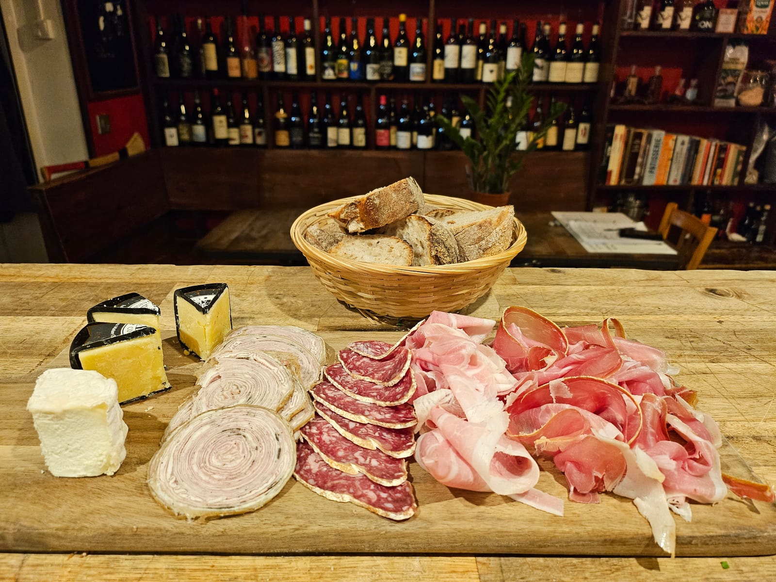Planche de fromages affinés et de charcuterie tranchée minute, panier de pain maison, devant les rayonnages de vins de la cave